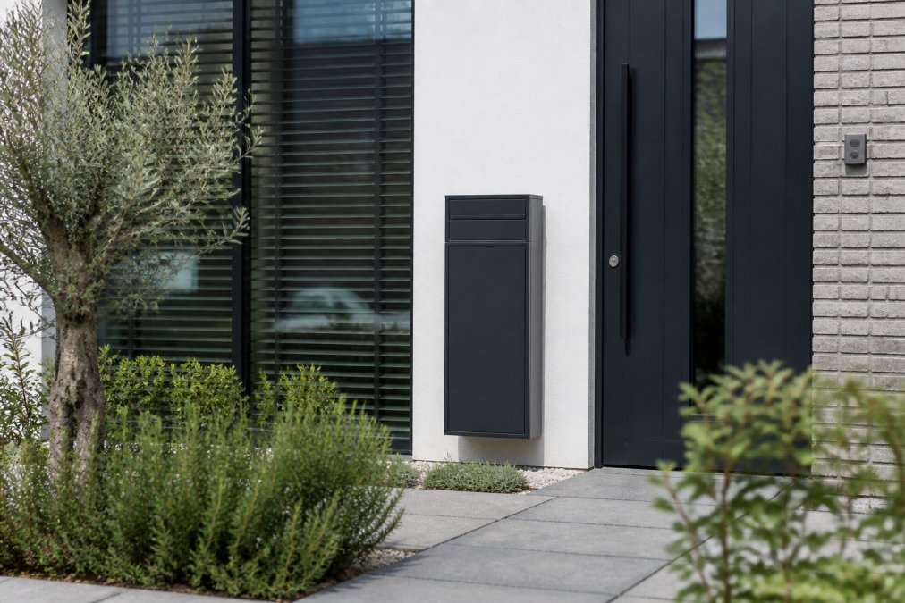 Strak antracietkleurige pakketbrievenbus gemonteerd op de gevel van een moderne Nederlandse woning, omgeven door een verzorgde kleine voortuin met klinkers en lage heesters, fotografisch vastgelegd in zacht daglicht