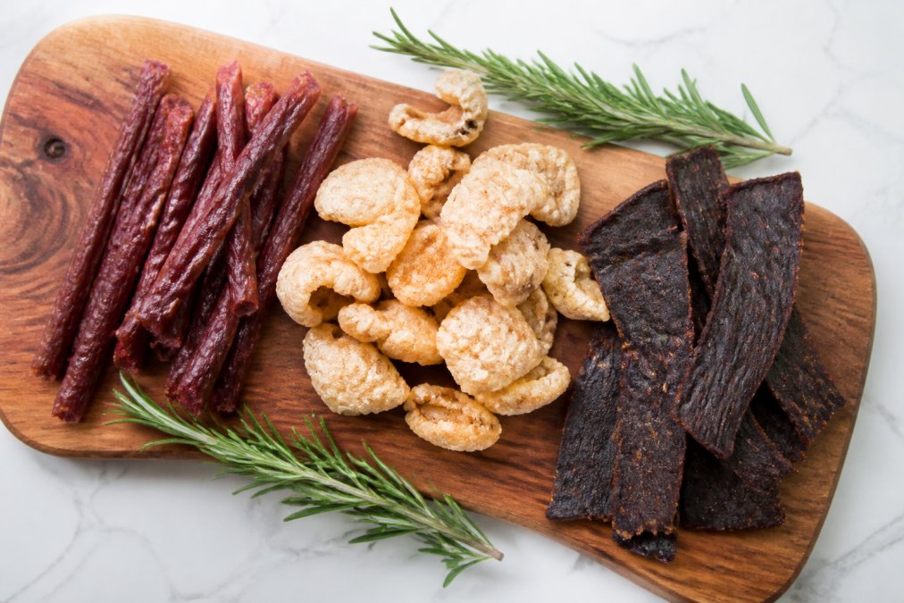 Overhead shot of grass-fed beef sticks, pork rinds, and jerky arranged on a rustic wooden board with fresh rosemary sprigs and a clean white background, bright natural lighting