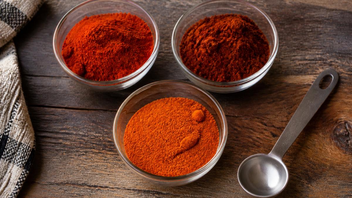 Overhead shot of small glass bowls filled with different red spice powders (paprika, chili powder, cayenne) arranged on a rustic wooden kitchen counter, with a measuring spoon resting beside them