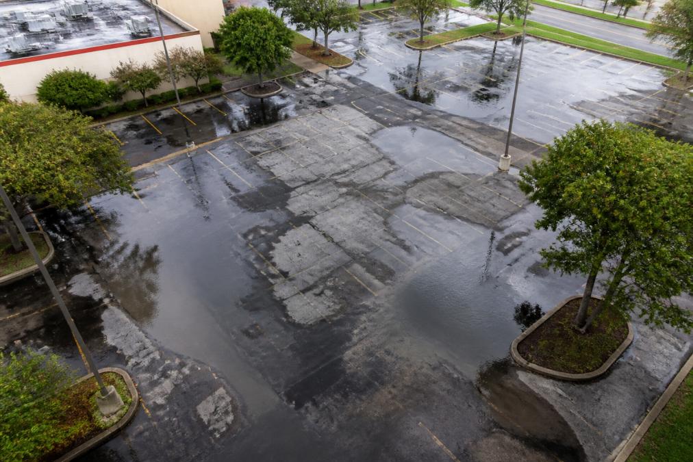 Aerial view of commercial parking lot with visible standing water pooling in multiple areas after heavy rain, showing wet patches and asphalt surface degradation