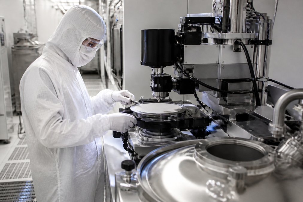 Semiconductor fabrication cleanroom environment with technician in full protective suit and hood working at precision metrology equipment station near wafer processing chamber, controlled lighting highlighting the instrumentation detail
