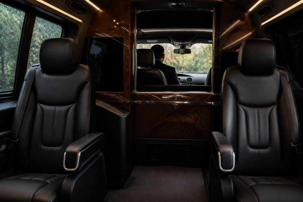 Interior view of a luxury van with premium leather captain's chairs, soft ambient lighting, and a professional chauffeur visible through a privacy partition, showing refined wood trim and climate control panel