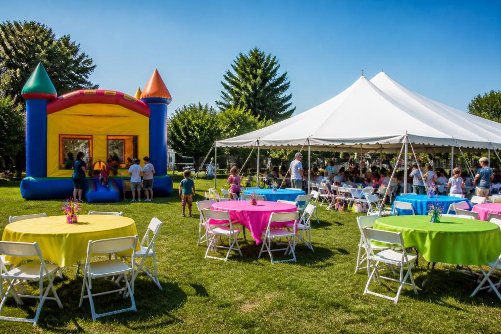 Colorful outdoor party setup with a bounce house, frame tent, tables, and chairs arranged for a children's birthday celebration, with families enjoying the event in the background under clear skies