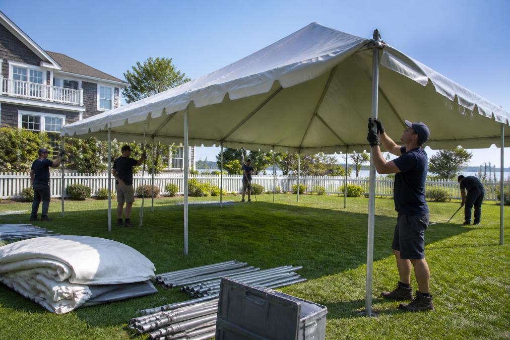 Professional event rental team setting up a frame tent on a residential lawn, with team members positioning poles and securing fabric under clear skies, demonstrating proper installation technique