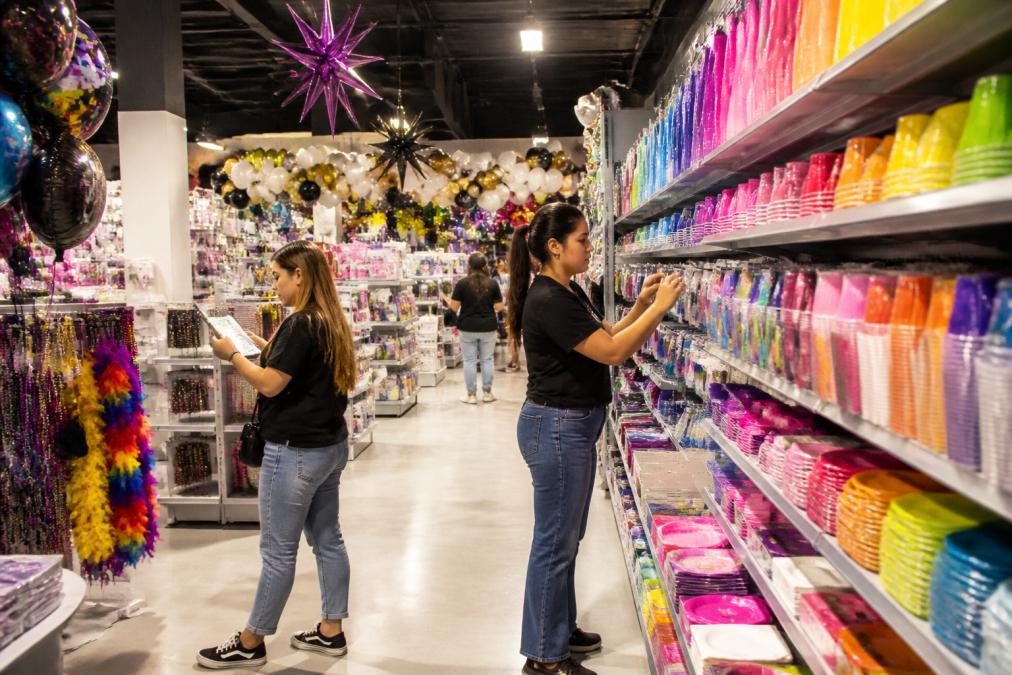 Interior of a vibrant party supply store with colorful shelves displaying hundreds of balloons, themed party kits, tableware sets, decorations, and costumes organized by color and occasion, with customers browsing and staff arranging displays
