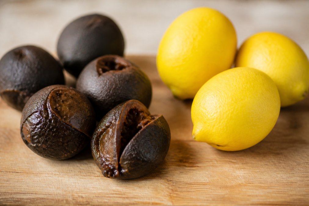 Close-up de citrons noirs fermentés et de citrons blancs frais côte à côte sur un plan de travail en bois, montrant le contraste de texture et de couleur sous une lumière naturelle douce