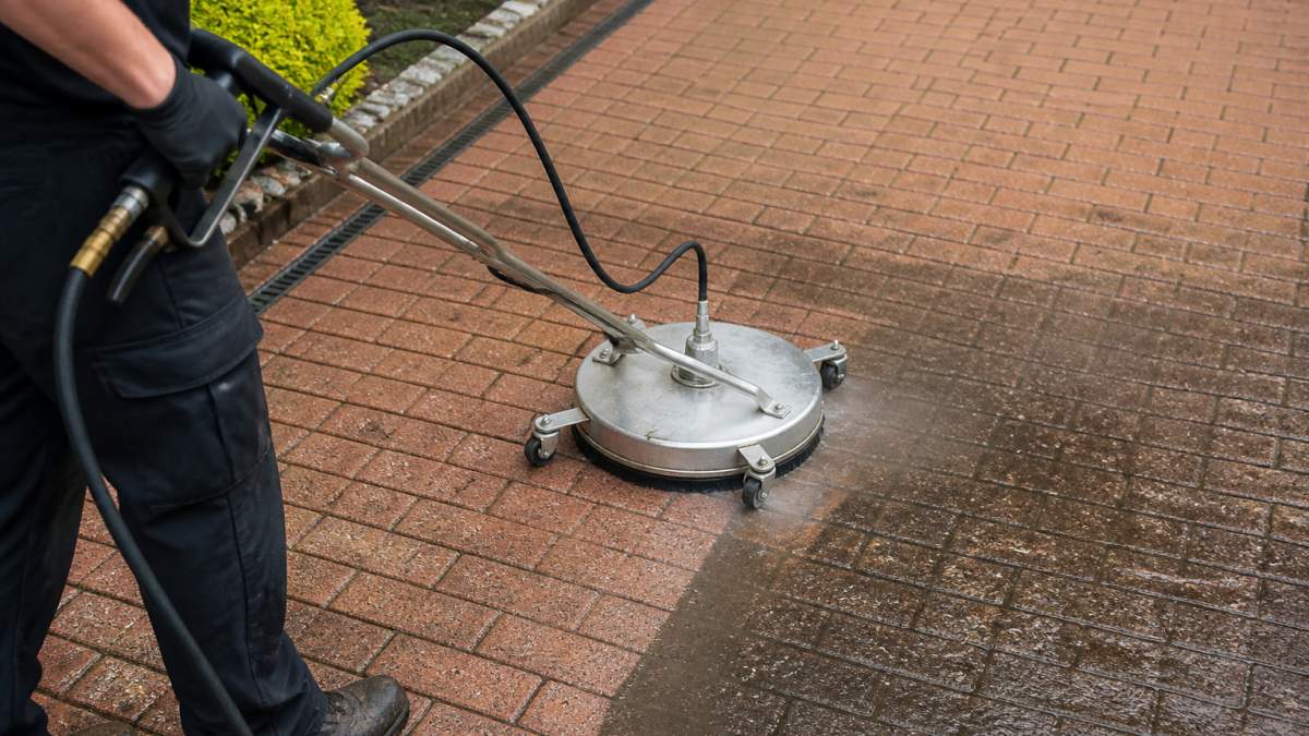 A professional cleaner using a rotary surface cleaner attachment on a block paving driveway, with water spray visible and a clean strip contrasting with the dirty surface
