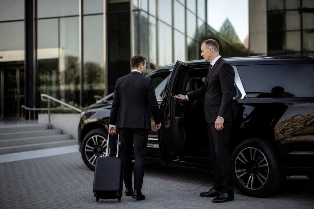 Professional driver in dark suit opening door of luxury black SUV for business executive with luggage at modern office building entrance, morning light