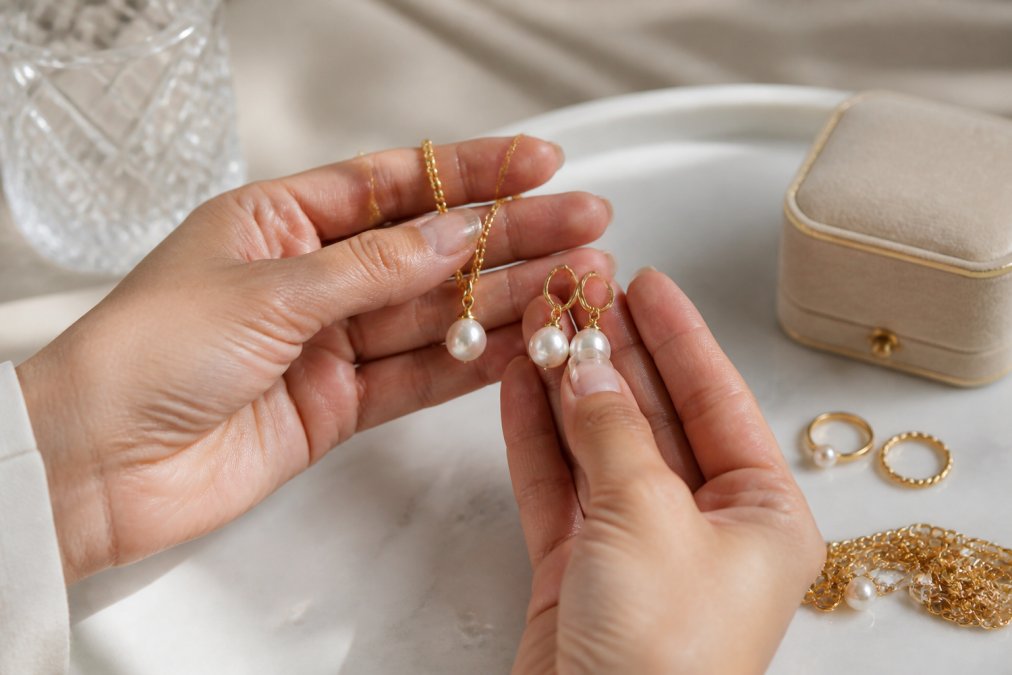 Close-up photograph of hands holding and examining freshwater pearl earrings and gold-plated necklace, showing the detailed lustre and texture of the pearls under natural lighting