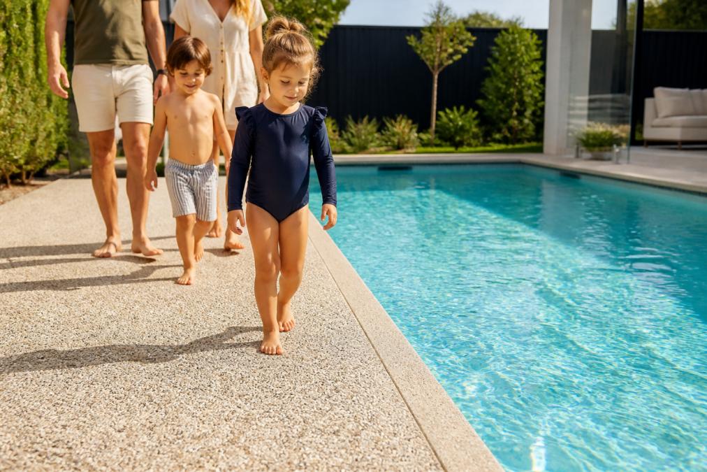 Family with young children walking barefoot on slip-resistant pool surrounds beside a sunny backyard swimming pool.