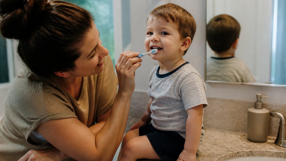 Parent gently brushing a toddler's teeth with a soft-bristled brush at a slight 45-degree angle, child sitting on bathroom counter with soft lighting, small smile on child's face Parent gently brushing a toddler's teeth with a soft-bristled brush at a slight 45-degree angle, child sitting on bathroom counter with soft lighting, small smile on child's face