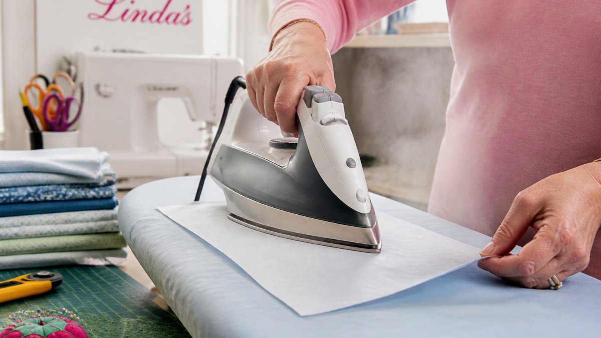 Hands using a hot iron to fuse white interfacing to the back of blue cotton fabric on an ironing board