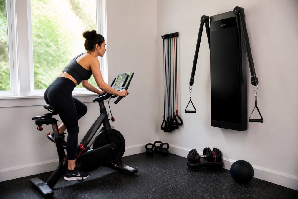 Person riding a compact indoor cycling bike near a wall-mounted strength trainer in a small home gym