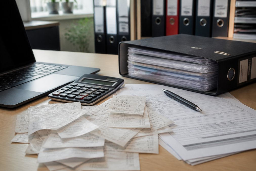 Arbeitsplatz mit aufgeklapptem Laptop, Taschenrechner, Quittungen und Ordner mit sortierten Dokumenten für die Steuererklärung unter Bürolicht
