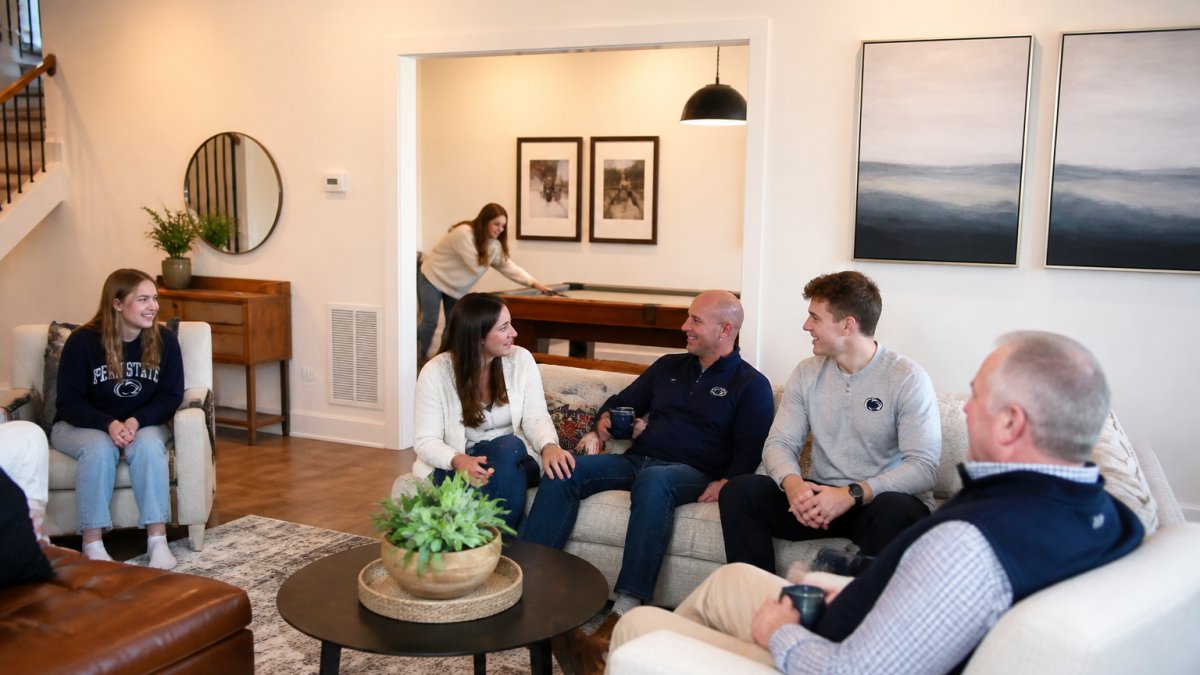 Family gathered in modern townhome living room with comfortable seating, game room visible in background, warm lighting, multiple generations relaxing together during Penn State weekend