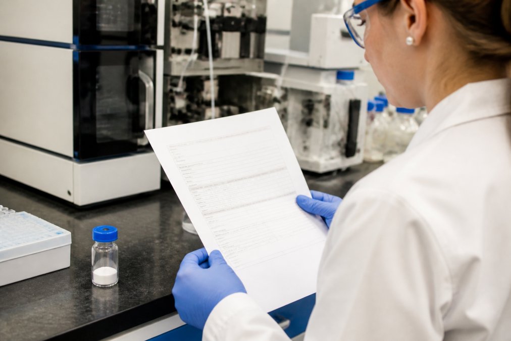 Researcher examining a printed Certificate of Analysis document at a laboratory bench, with a vial of lyophilized peptide and HPLC equipment visible in the background under bright laboratory lighting