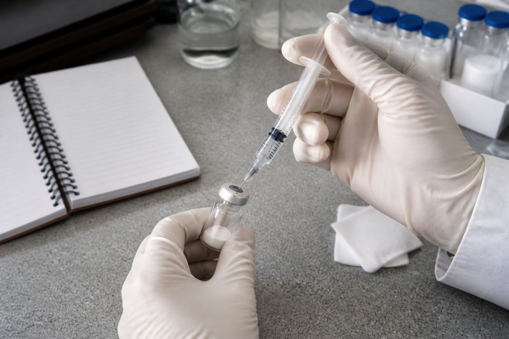 A researcher's gloved hands carefully injecting bacteriostatic water into a small glass peptide vial using a syringe, with a clean laboratory bench, alcohol swabs, and a notebook in the background