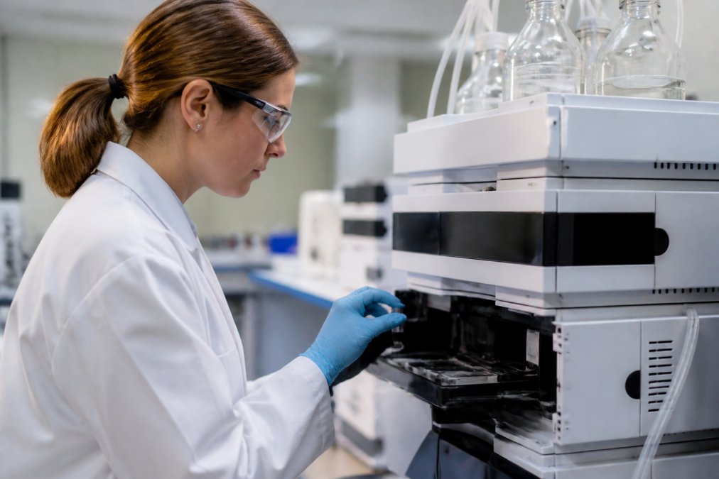 Laboratory technician in white coat and gloves carefully inserting a peptide sample vial into an HPLC instrument in a modern analytical chemistry lab with focused task lighting