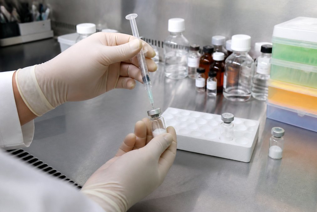 Researcher's hands carefully reconstituting a lyophilized peptide vial with bacteriostatic water using a sterile syringe and needle under controlled laboratory conditions, with storage containers and labels visible in the background