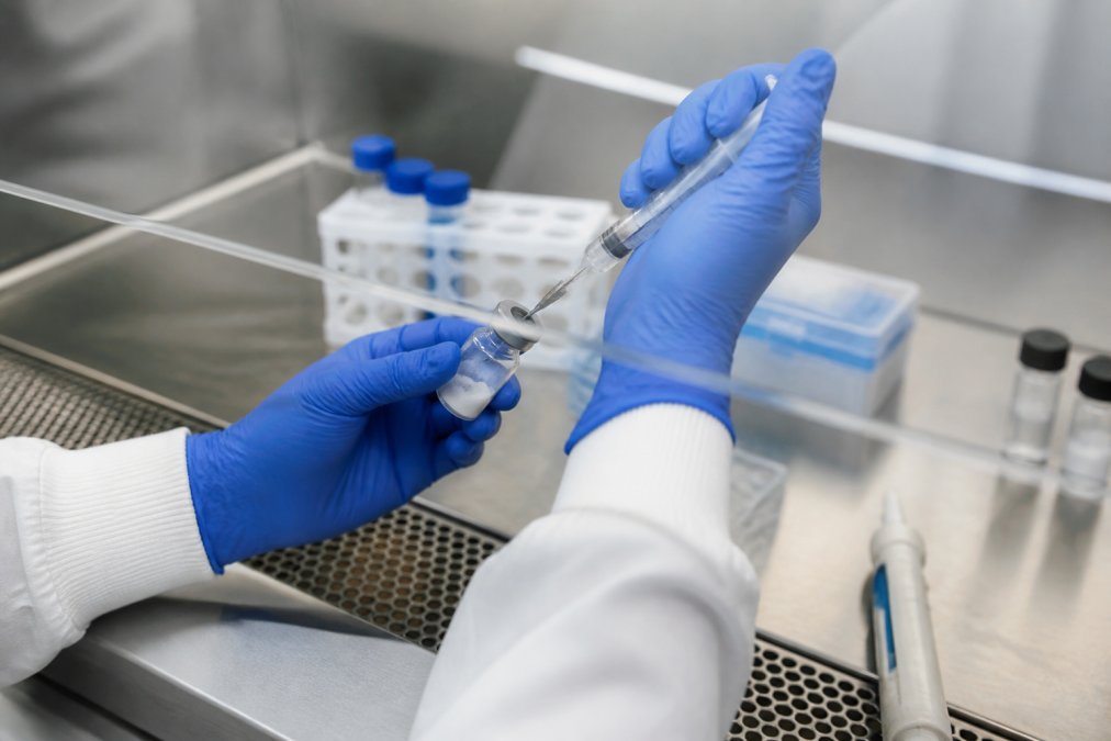 Close-up of hands in a laminar flow hood carefully reconstituting a lyophilized peptide vial with bacteriostatic water using a sterile syringe, showing proper aseptic technique with clear focus on needle insertion angle and vial positioning
