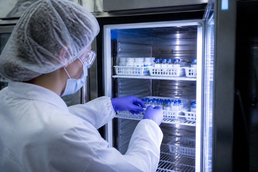 Laboratory technician in protective equipment carefully arranging peptide vials in a temperature-controlled storage unit with organized labeling and documentation visible on the shelf, cool blue lighting reflecting off the equipment