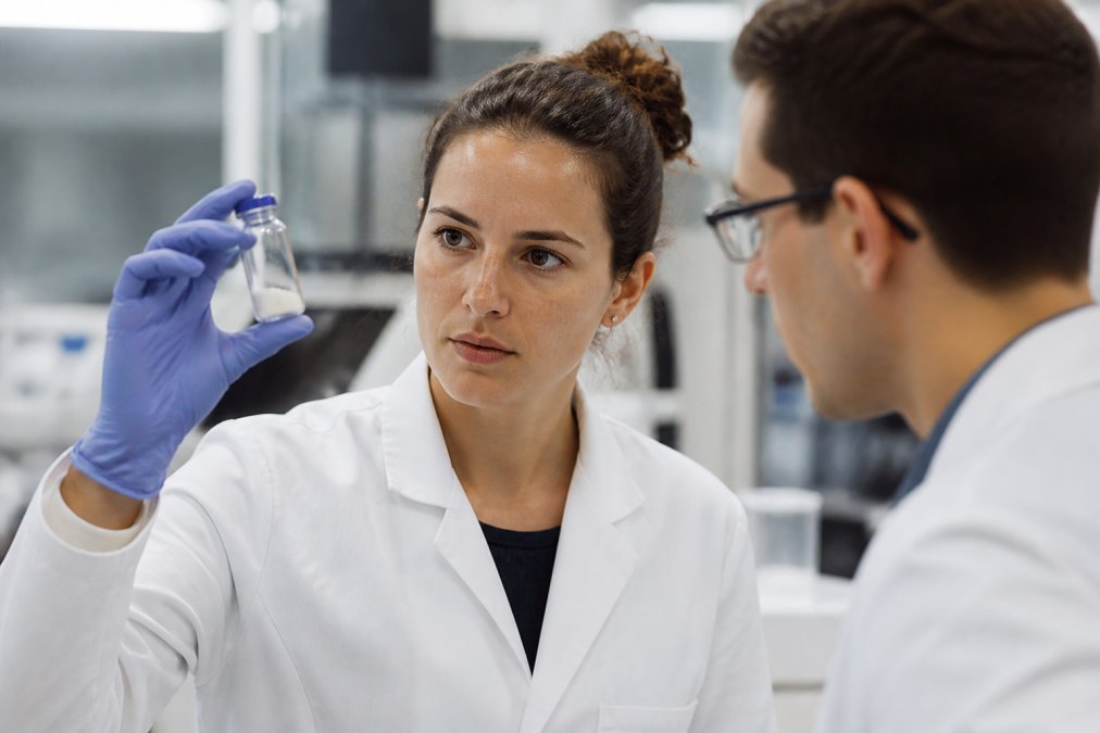 Research scientist in white lab coat inspecting a clear glass vial of peptide powder under bright laboratory lighting, with analytical equipment visible in the background