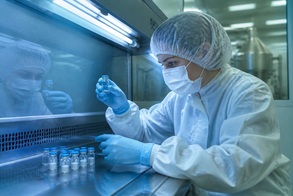 Laboratoriummedewerker in steriele schone ruimte met beschermende kleding en handschoenen voert kwaliteitscontrole uit op peptideflessen onder fluorescentverlichting