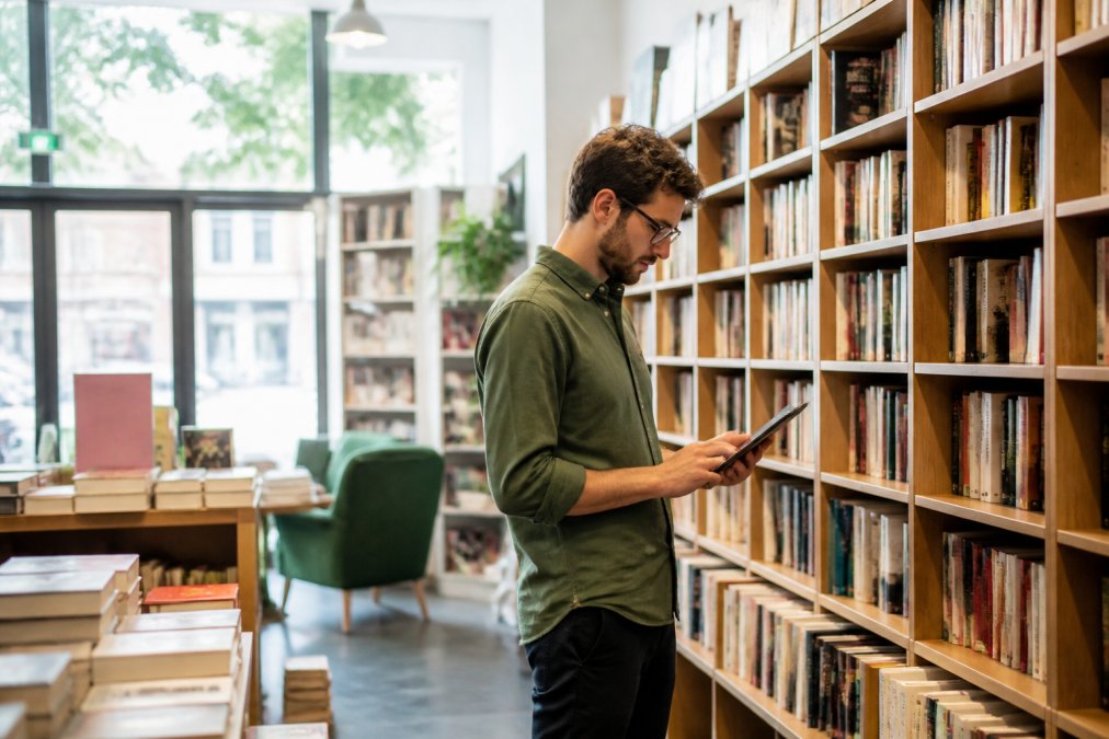 Un libraio che consulta un tablet con i dati di magazzino tra gli scaffali di una libreria indipendente ben organizzata, luce naturale dalla vetrina, atmosfera accogliente