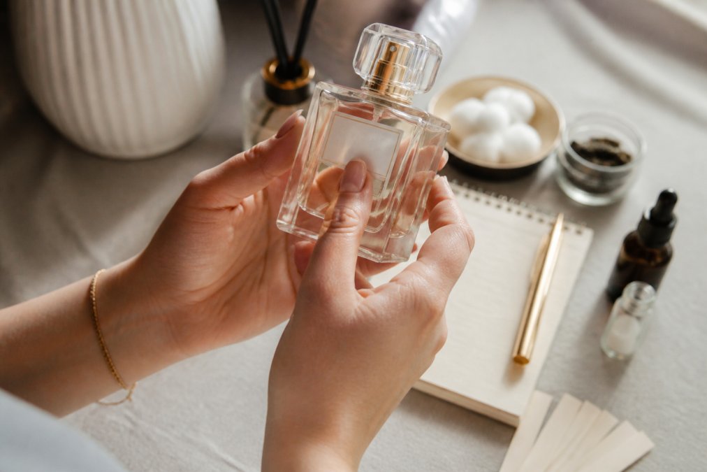 Close-up of hands holding a clear perfume bottle, examining the ingredient label with natural window lighting illuminating the text, neutral background