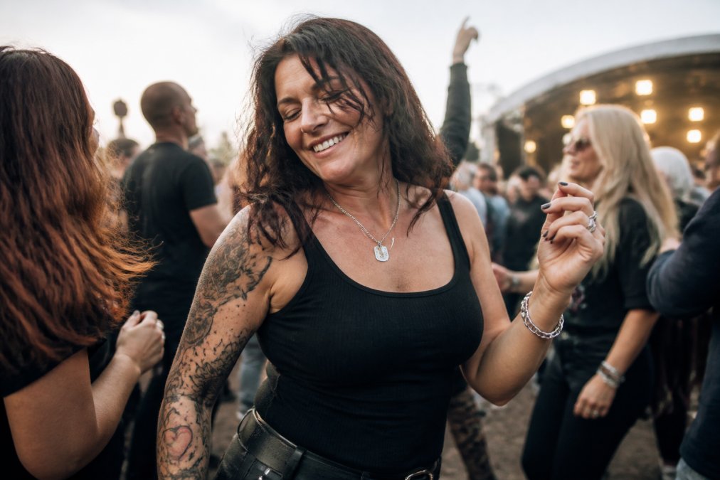 Woman in her 40s at a live outdoor music venue, dancing with friends in genuine joy, warm evening light, relaxed shoulders, real smile, surrounded by other people enjoying the moment