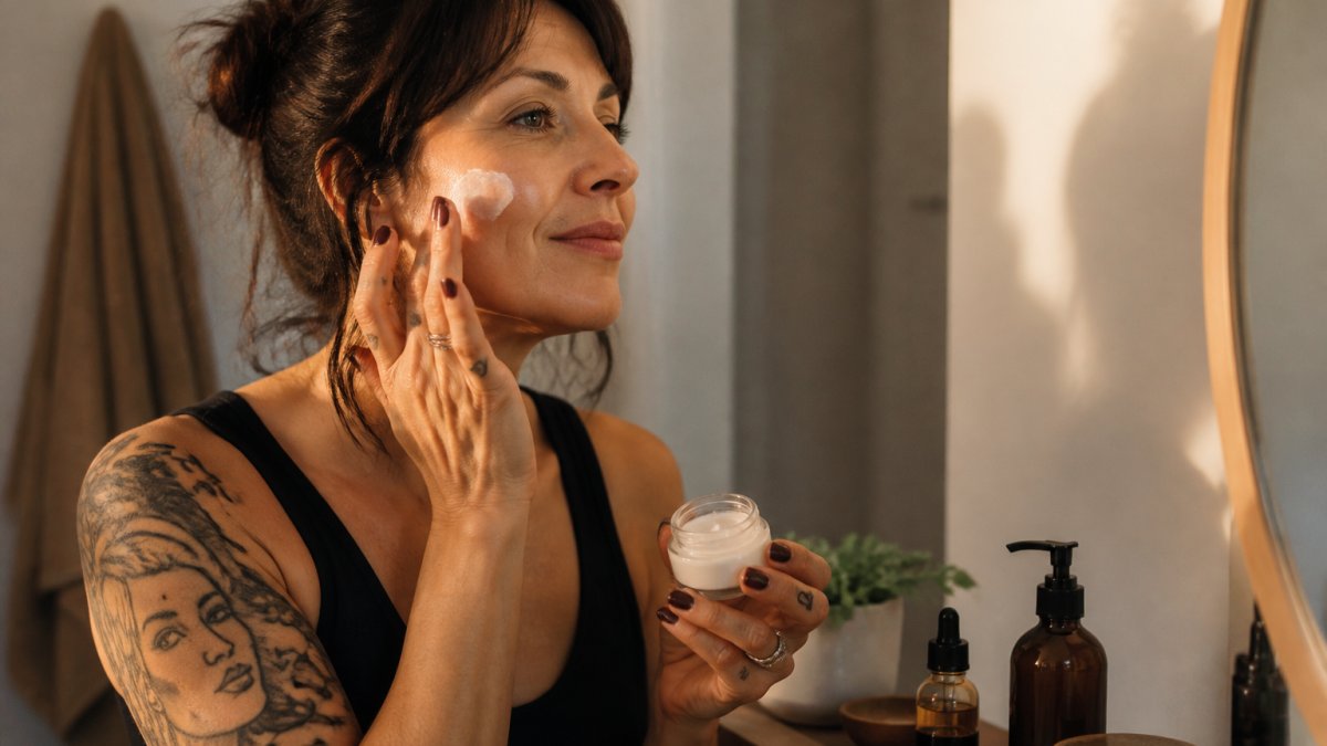 Woman in her 40s applying skincare product to her face in natural morning light, looking relaxed and confident