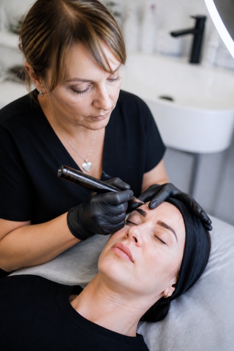 Permanent makeup artist using a microblading tool on a client's brow during a session influencing touch up pricing