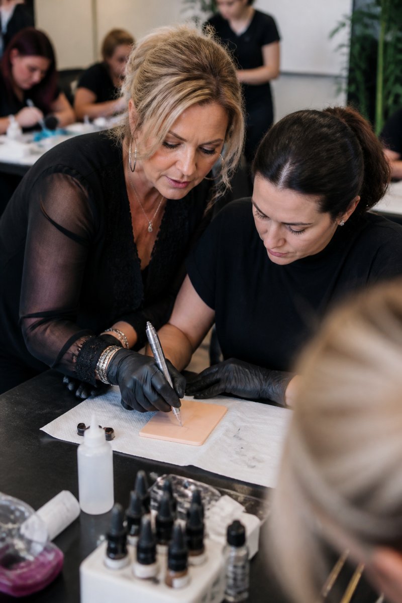 A master instructor guiding a student's hand during a practice session on a synthetic skin pad in a bright, professional training studio