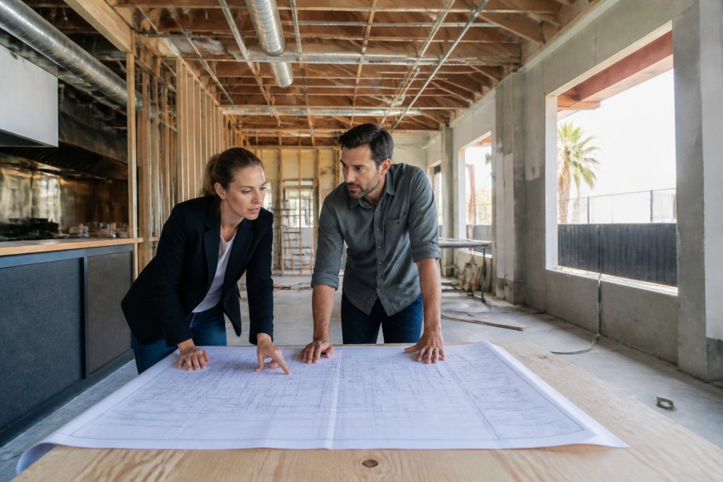 An architect and a restaurant owner leaning over large-scale permit drawings spread across a plywood table inside a restaurant under construction, exposed framing and conduit overhead, natural light through unglazed window openings