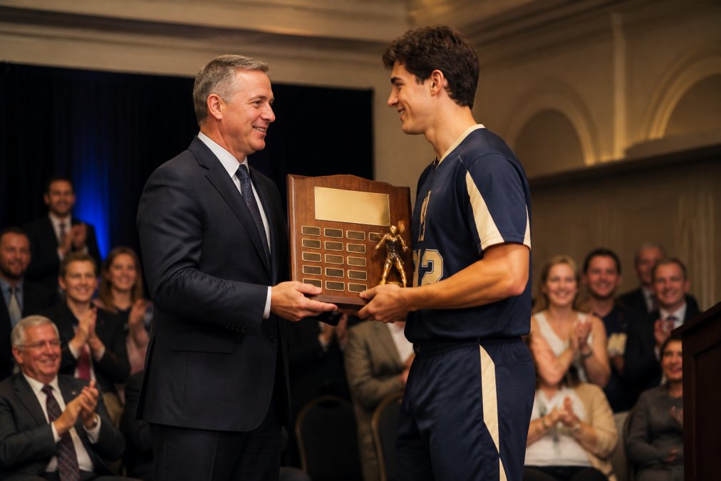 Athletic director presenting a perpetual trophy to a team captain at an award ceremony, with audience members watching and applauding in the background, formal event lighting