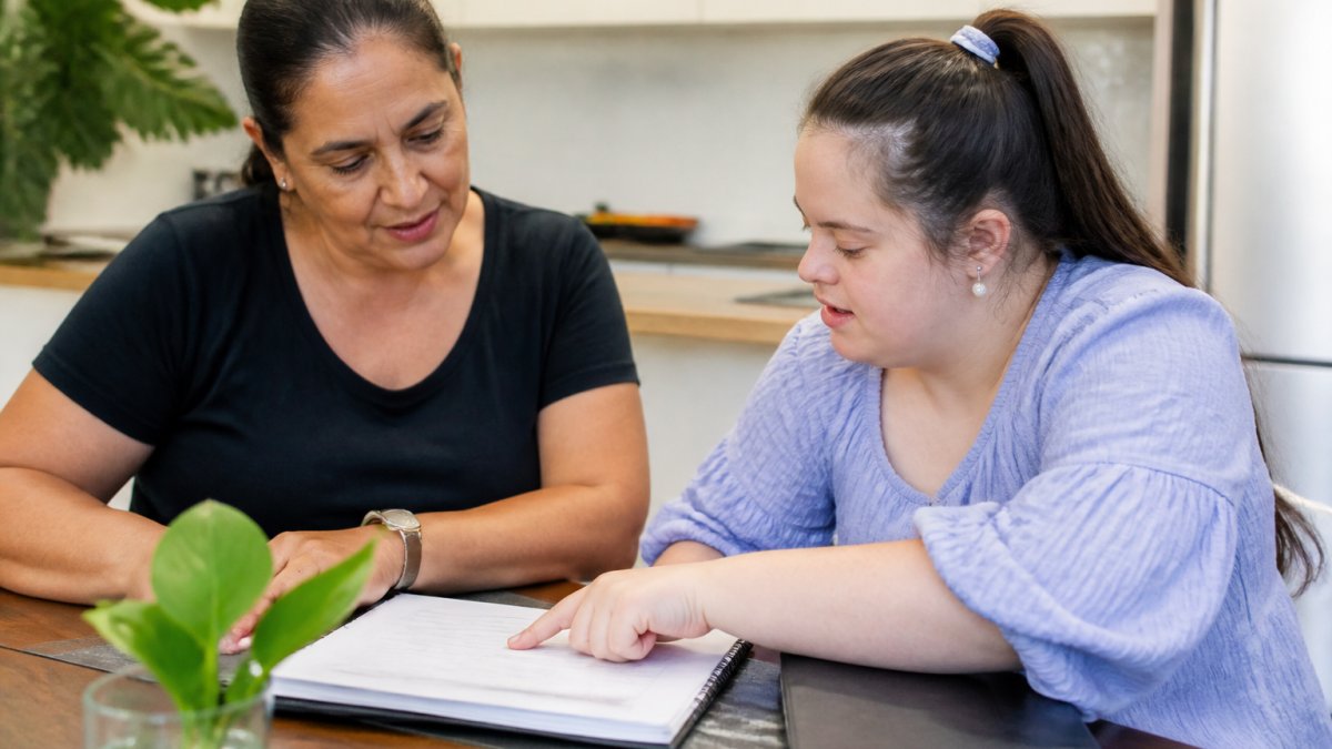 Professional support worker and NDIS participant reviewing a care plan document together at a table, with the participant pointing to specific goals written on the page, natural office lighting
