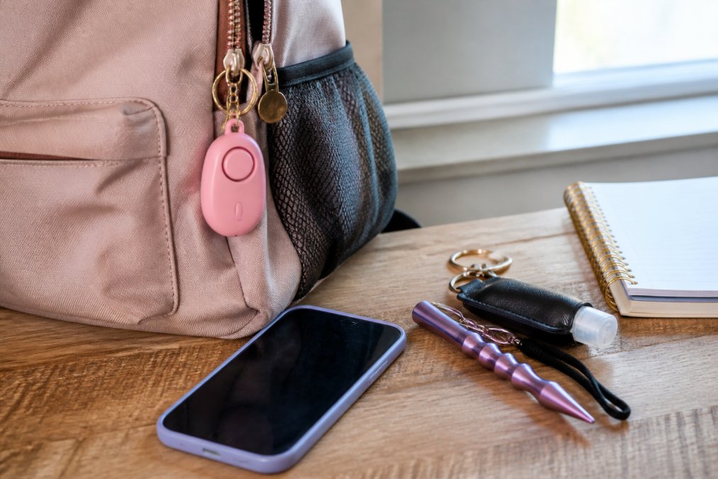A close-up of a college student's backpack with personal safety items visible: a keychain alarm attached to the bag strap, a phone with a safety app open on the desk next to it, and a compact self-defense tool arranged on a wooden desk, natural window lighting