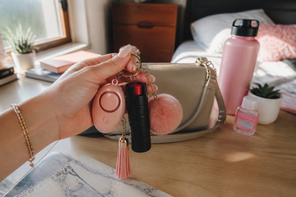 Close-up of a young woman's hand holding a keychain set with a personal alarm, small pepper spray canister, and stylish accessories laid out on a college dorm desk with soft afternoon light coming through a window