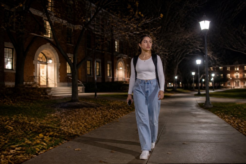 A college-age woman walking alone at night on a well-lit campus path, looking alert and holding her phone at her side, with brick campus buildings and streetlamps visible in the background