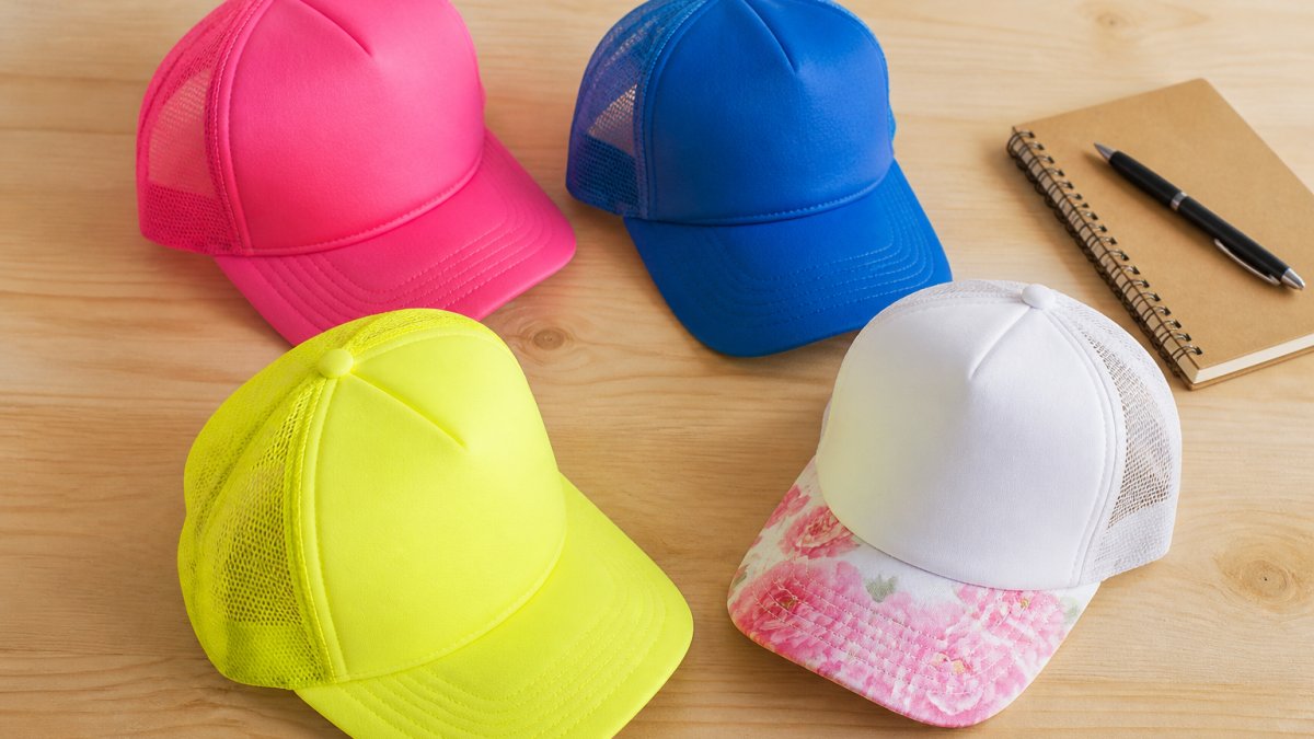 Flat lay of four trucker hats in neon pink, blue, bright yellow, and white with a pink peony print bill, arranged on a light wood table with a small notebook and pen nearby