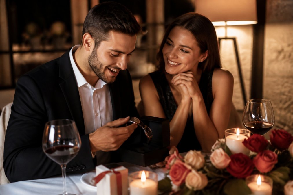A couple seated at a candlelit dining table, one partner lifting an engraved watch from a gift box while the other leans in with a warm smile, soft floral centerpiece and warm ambient light filling the background