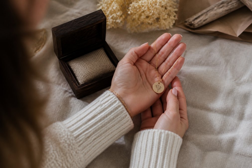 Close-up of a woman's hands holding a delicate engraved necklace and a small custom keepsake box on a soft linen surface, warm natural light