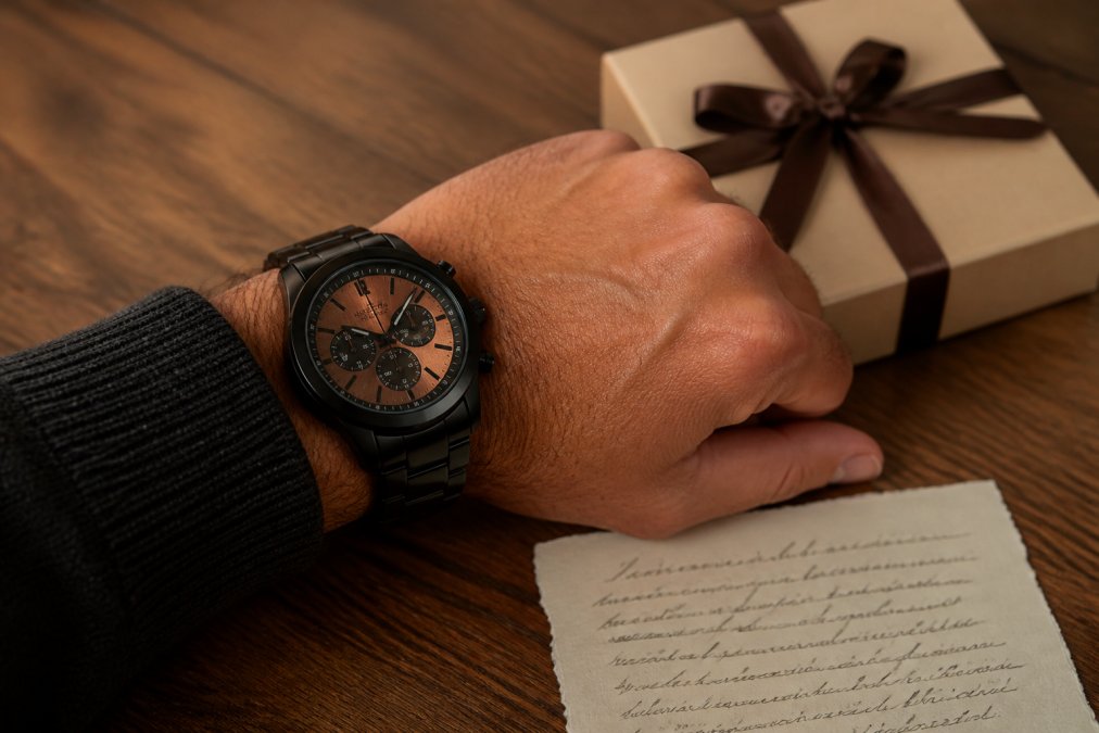 A close-up of a man's wrist wearing a black chronograph watch with a copper dial, resting on a wooden table next to a small gift box and a handwritten note