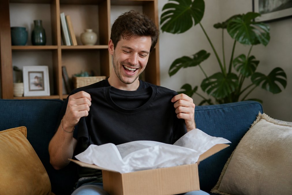 A man in his late 20s wearing a black novelty t-shirt with a funny brother slogan, laughing while opening a gift box in a casual living room setting