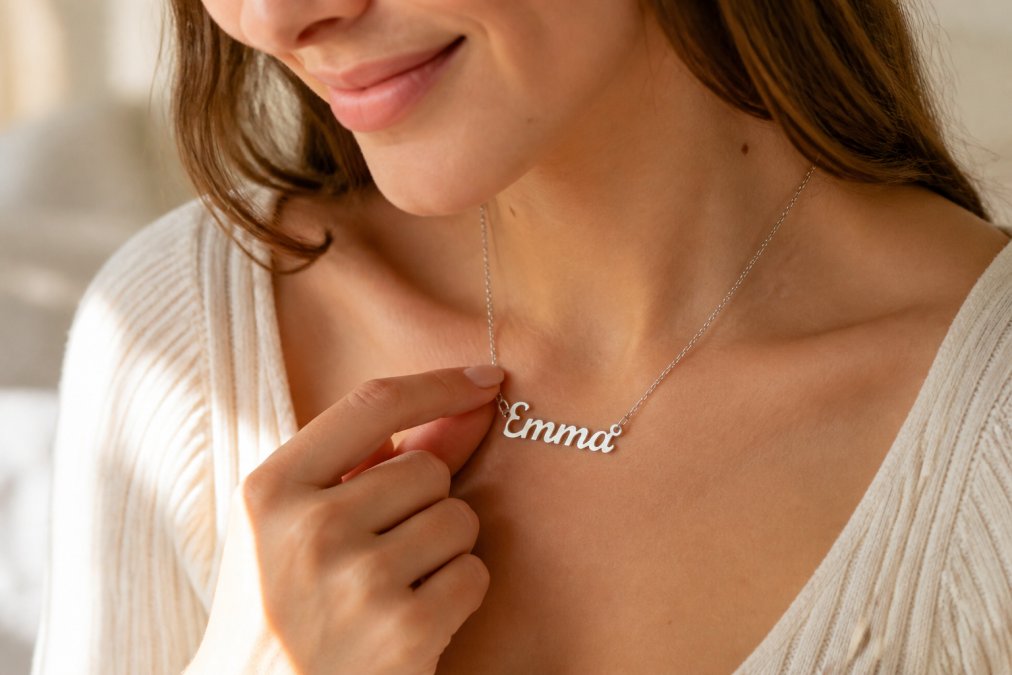 Woman wearing an elegant personalized name necklace, smiling softly while touching the pendant with natural lighting and warm tones showing how the piece sits on her collarbone