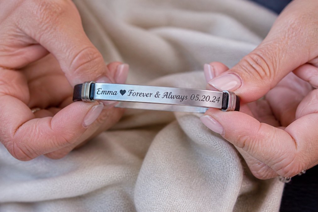 Close-up of hands carefully holding an engraved stainless steel bracelet, the custom lettering and date clearly visible in natural light, showing the fine detail of the craftsmanship and personalization