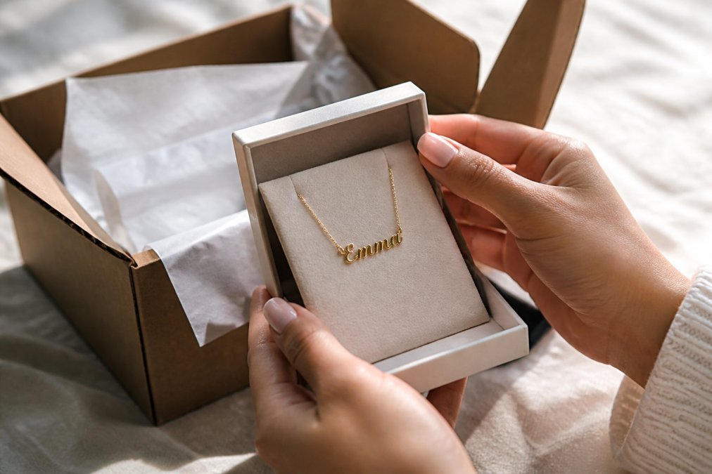 Close-up of a woman's hands holding a delicate gold name necklace in a soft-touch gift box, with natural light highlighting the cursive engraving