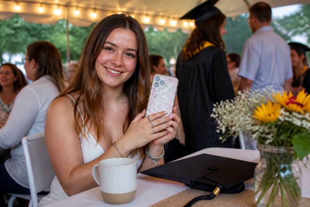 A young woman holding a personalized floral phone case with her name on it, smiling while sitting at a graduation celebration table with a custom mug nearby