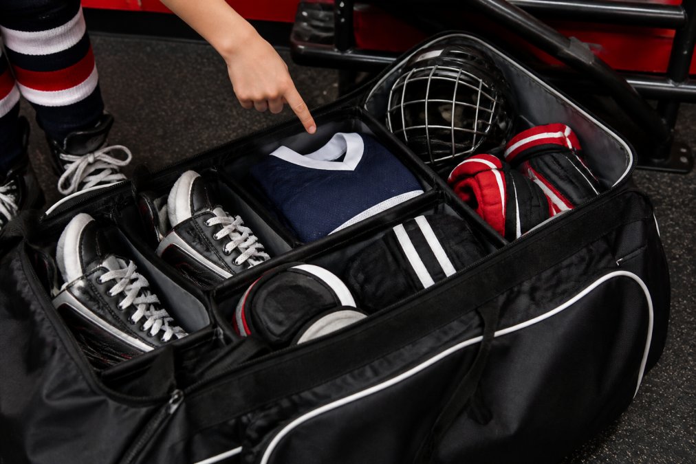 Open hockey equipment bag with organized gear neatly arranged inside, showing compartments filled with skates, gloves, helmet, and jersey, with a young hockey player's hand pointing to different sections of the bag