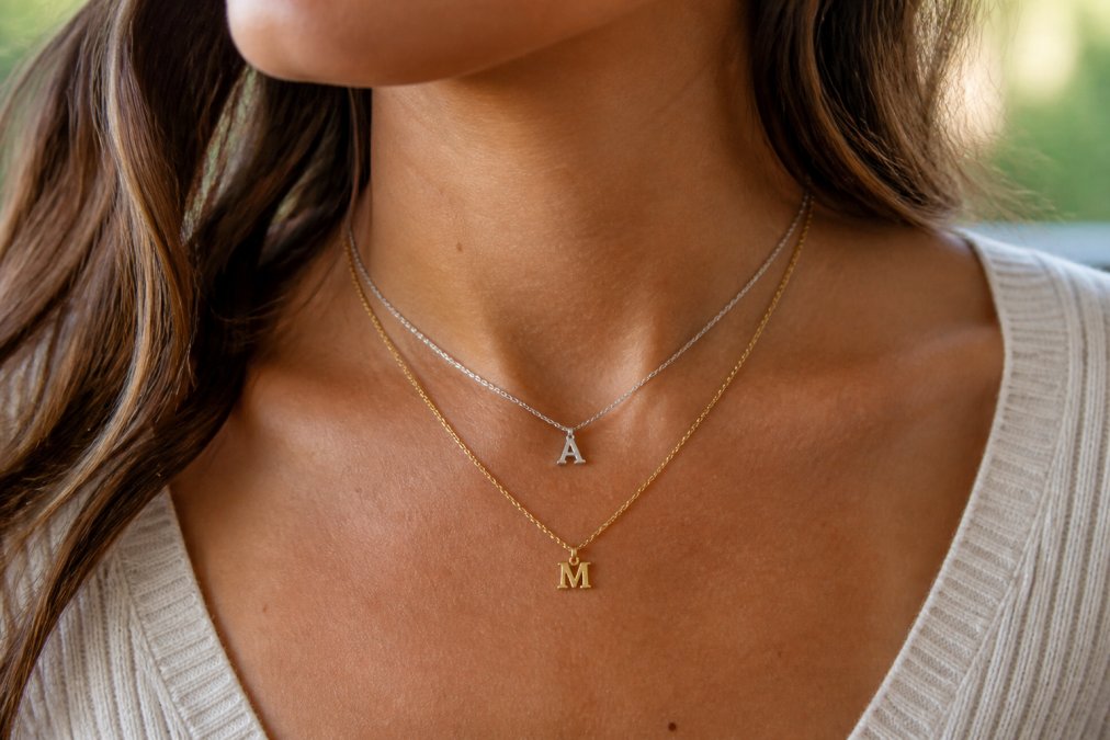 Close-up on a woman's neck and collarbone showing two layered initial necklaces, one gold and one silver, against a soft-focus neutral background with warm natural window light